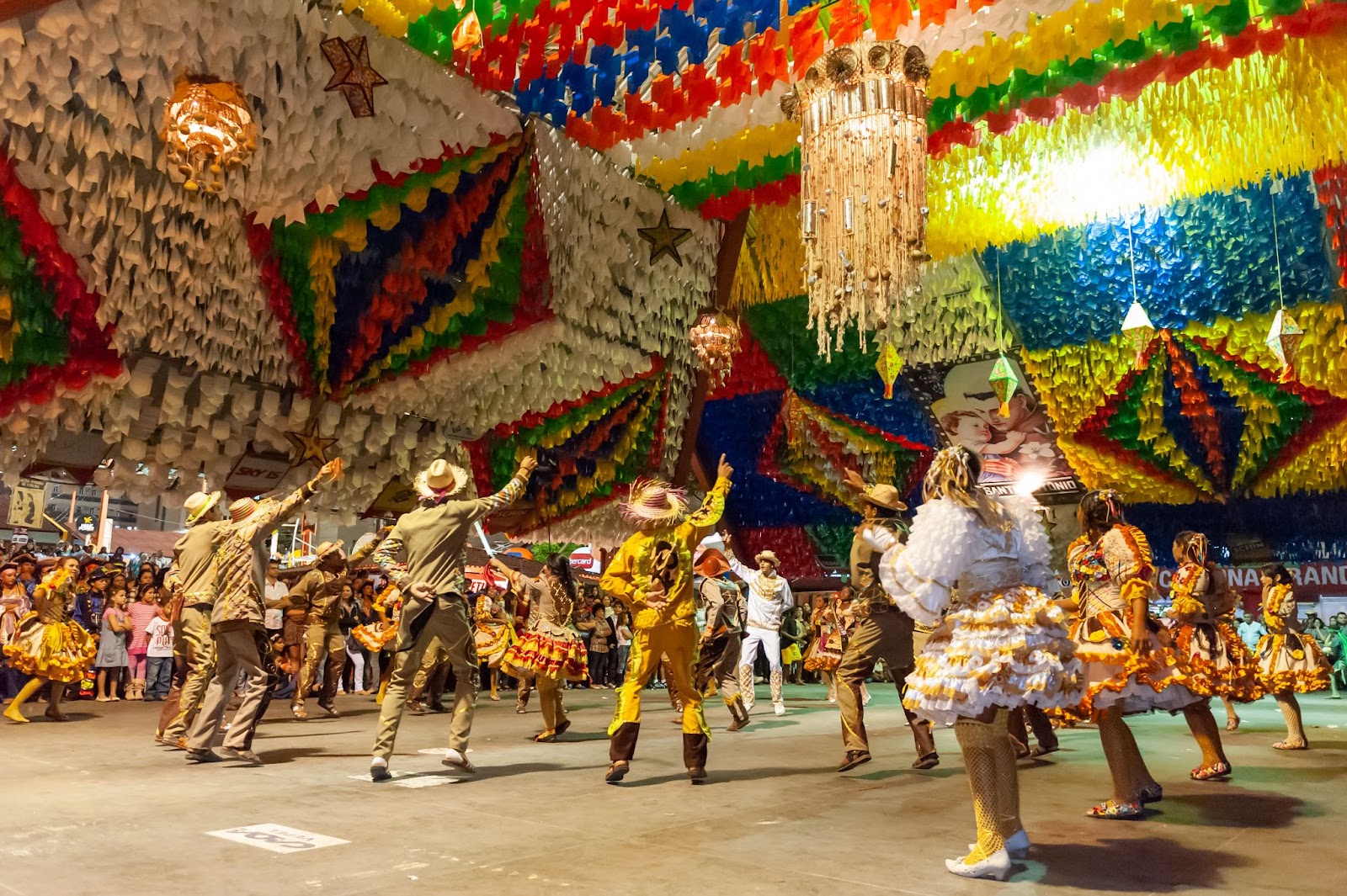 Quadrilha no São João de Campina Grande. Usando vestidos rodados e chapéus coloridos, os participantes dançam em roda na Praça do Povo. O teto está coberto por bandeiras coloridas e balões decorativos.