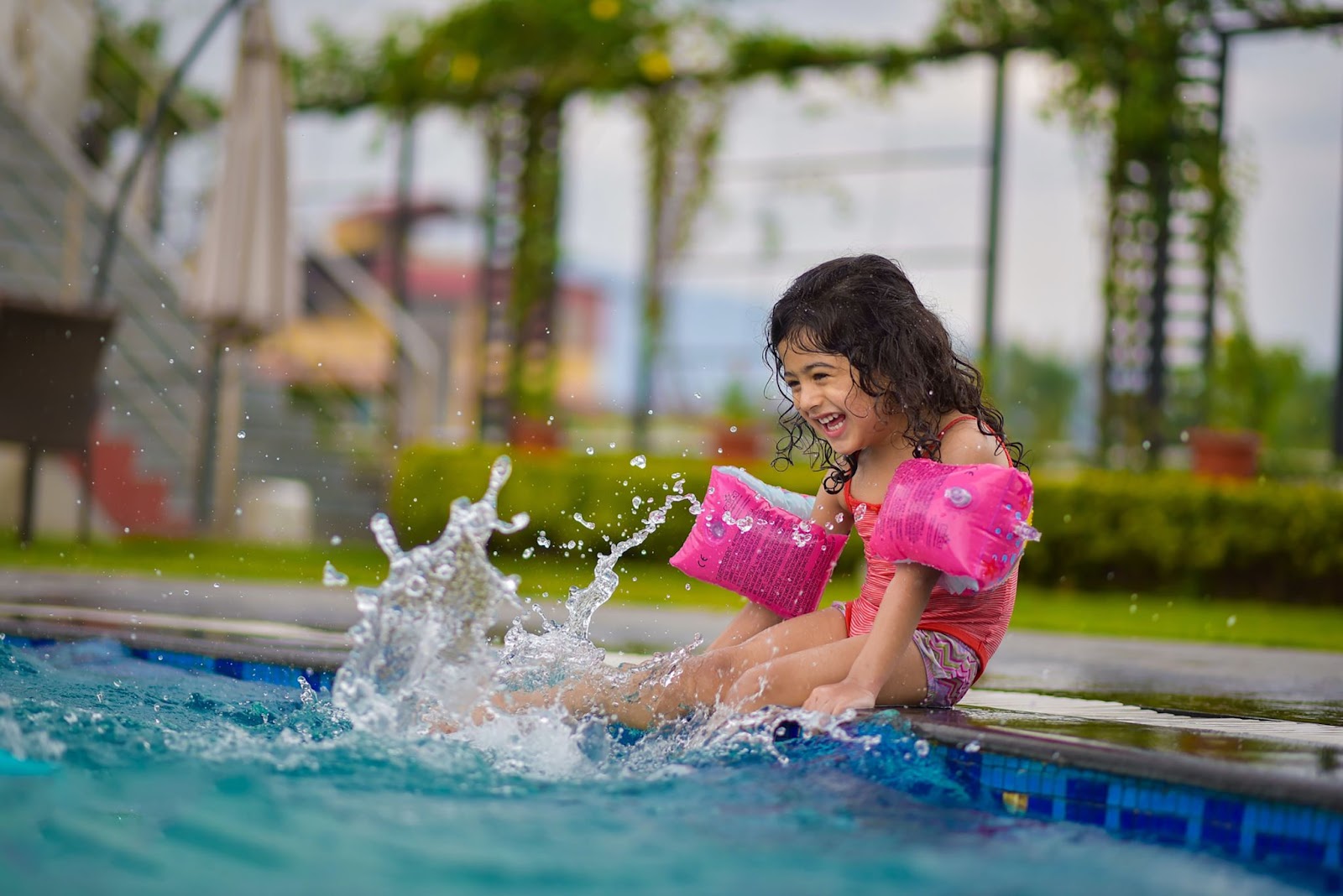 small girl with pink water wings kicking her feet by the side of a swimming pool