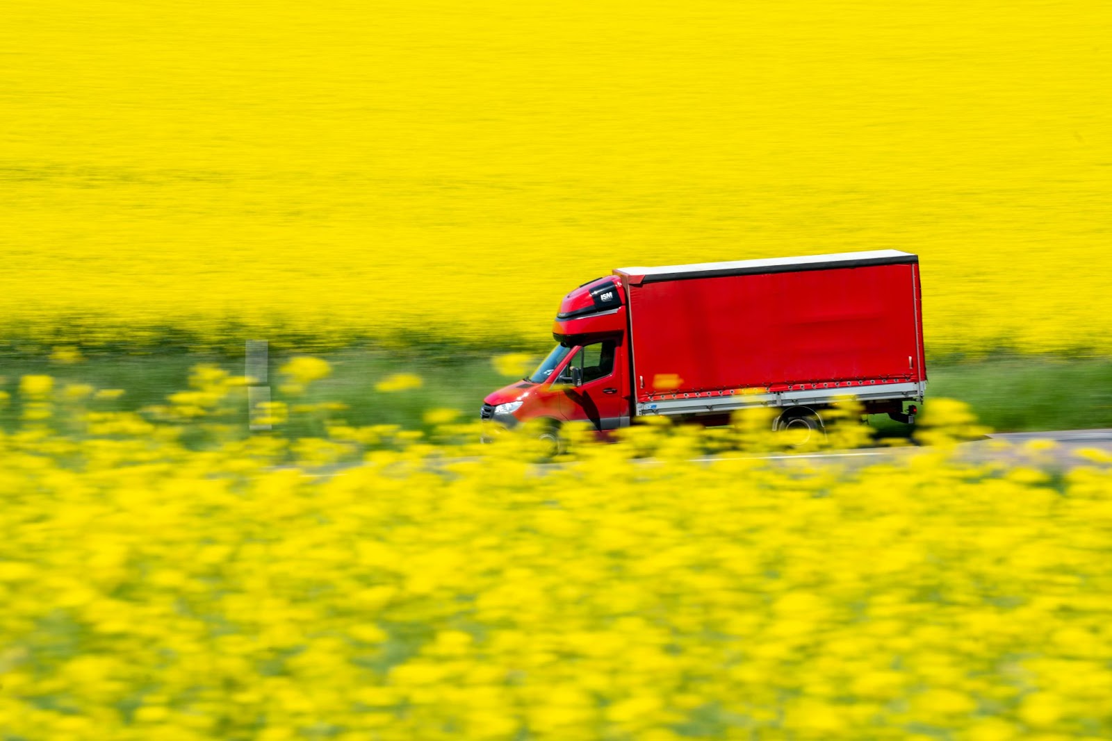 A red box truck driving on a road next to a field of yellow flowers