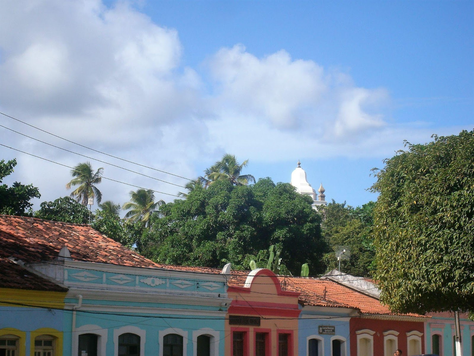 Foto que mostra o topo de casarões coloniais coloridos em Igarassu, com árvores atrás das casas e um céu azul com nuvens acinzentadas