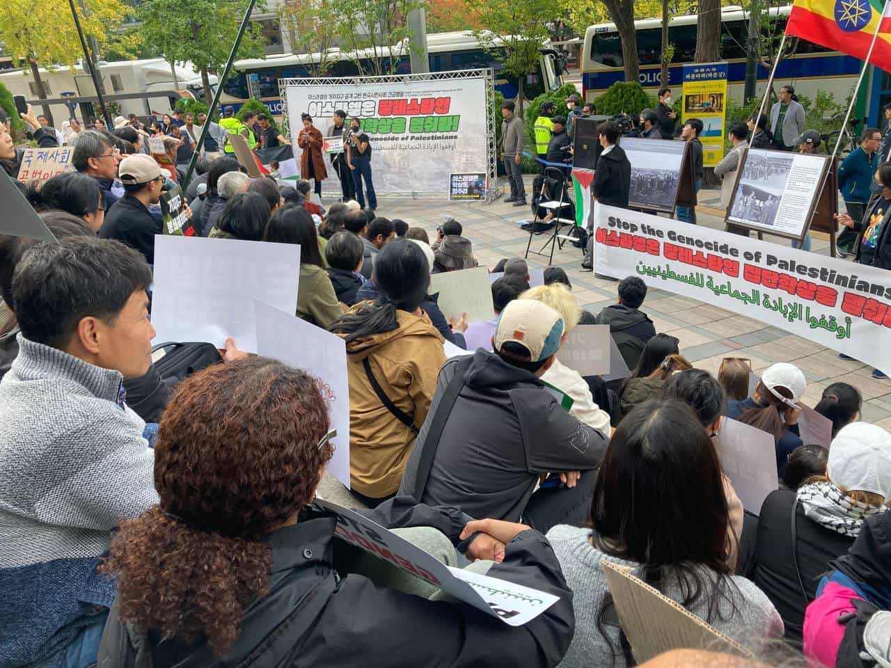 A crowd sit in a public square listening to a speech.