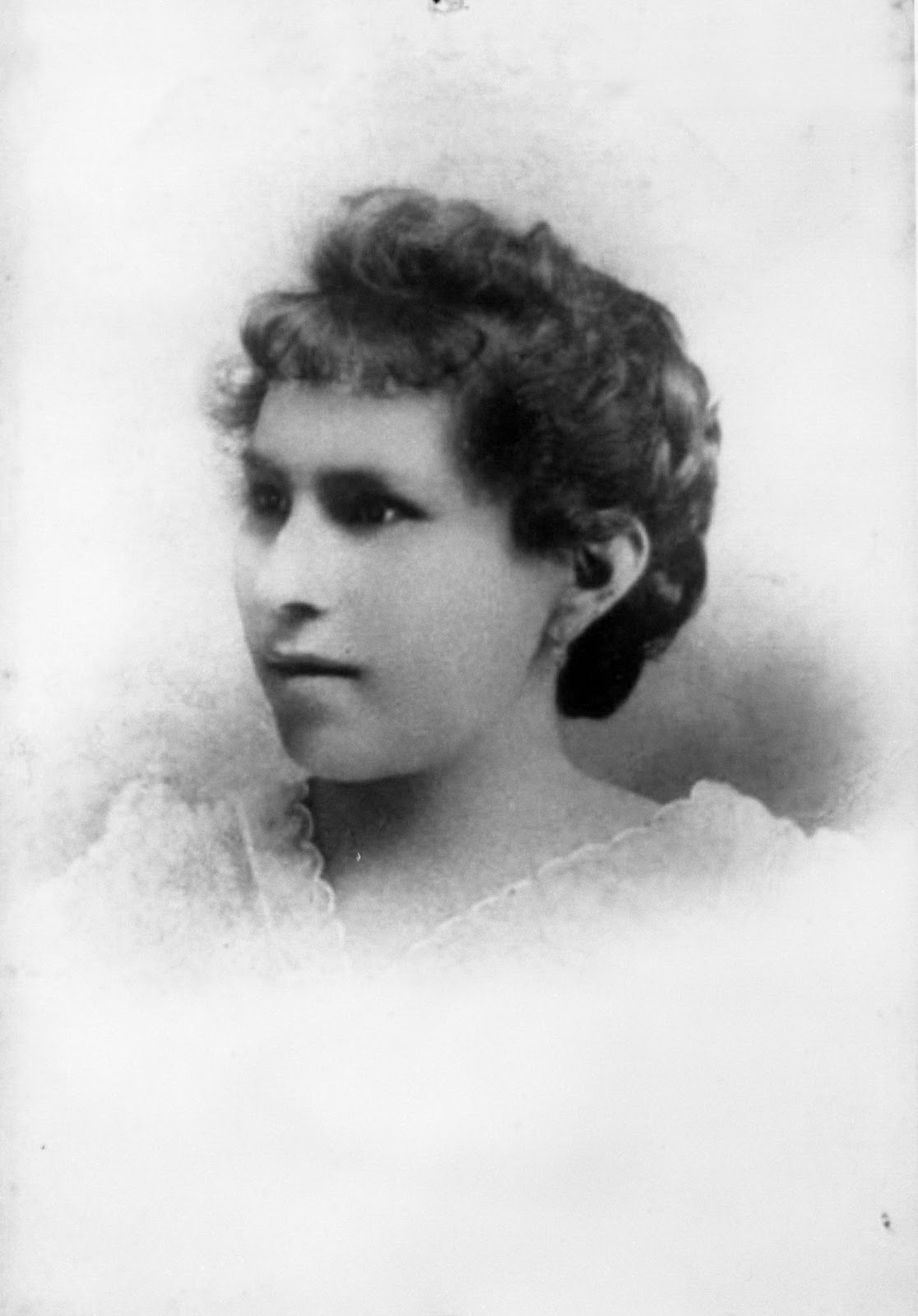 Rachael is looking to the left of the camera.  She has dark eyes and dark hair.  Her hair has short, curly bangs and is up in a bun.  She is wearing a white v-neck shirt with scalloped edging. The image has an oval vignette border fading as you move further out from the center.
