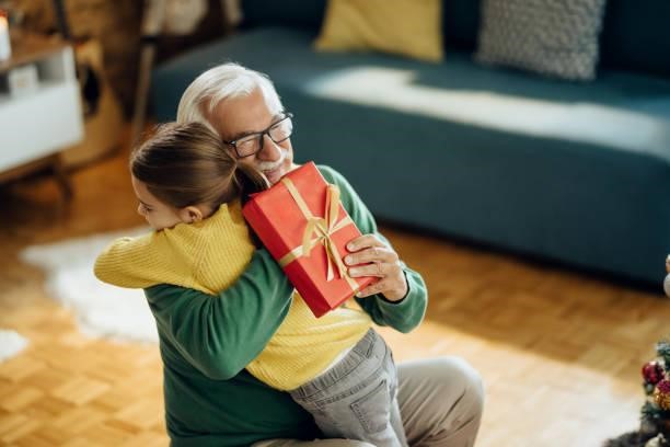 Grateful little girl embracing her grandfather while receiving Christmas present at home. Happy senior man embracing his granddaughter while receiving a gift on Christmas at home. Family Gift stock pictures, royalty-free photos & images