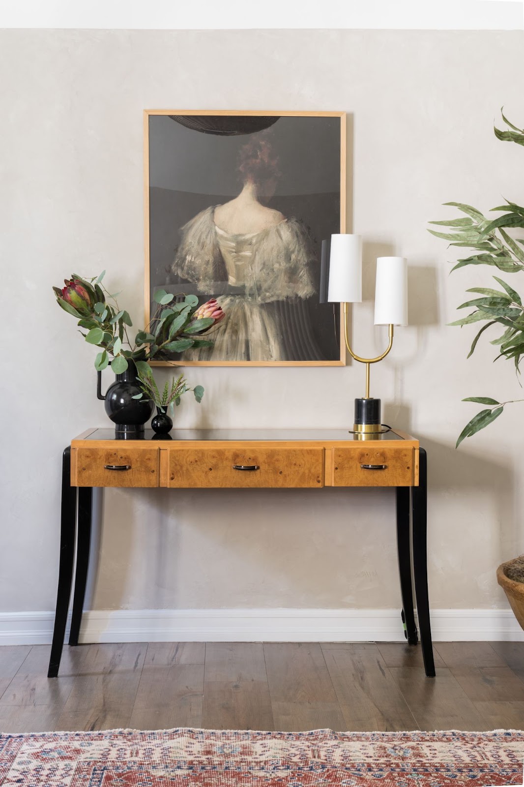Minimalist vignette: Neutral tones, a painting of a woman in a vintage dress facing away, a petite console table, black vases with floral branches, and a modern table lamp for understated elegance.