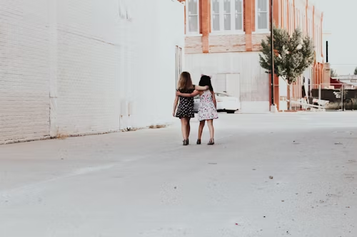 Two friends walking in the street side by side with their arms around each other. 