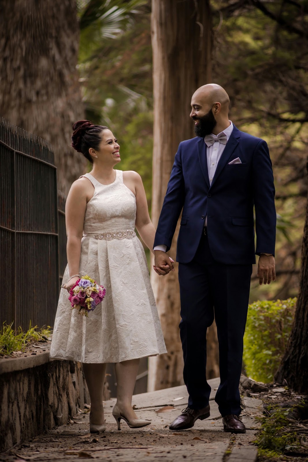 A bride holding hands with her groom, showcasing a short beaded waist gown with crisscross details.