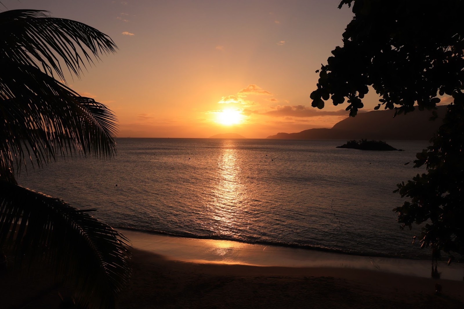 Praia com mar sem ondas ao entardecer, com o sol se pondo no horizonte, centralizado na imagem.