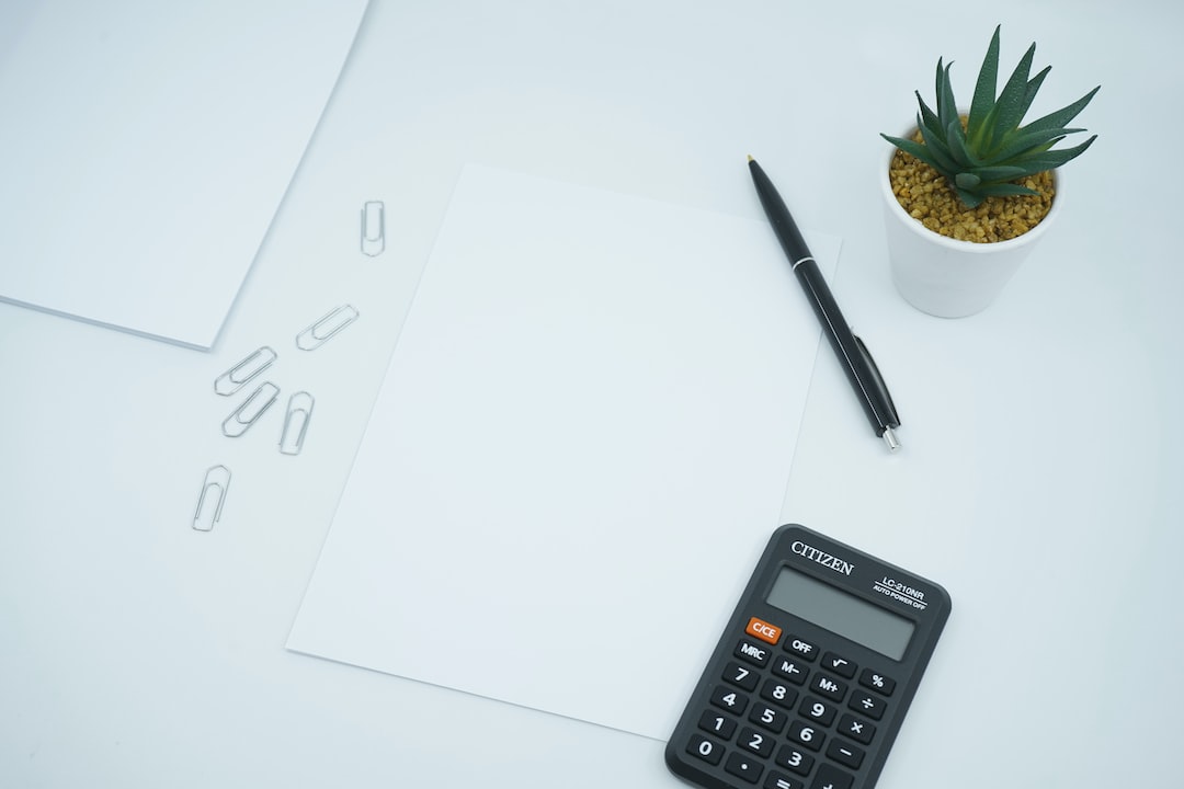 Een vel papier, een pen, een rekenmachine, wat paperclips, en een plant op een tafel. 