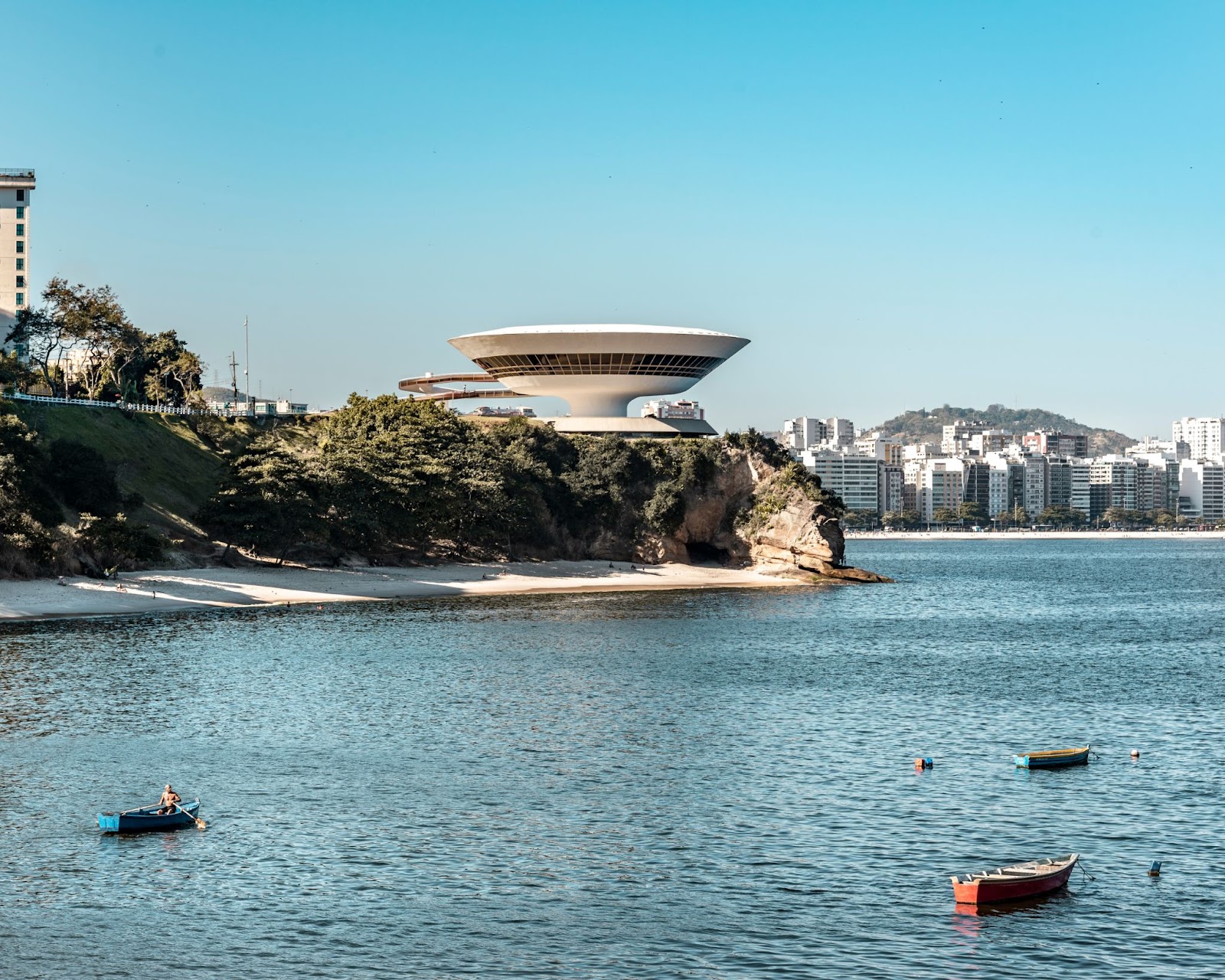 Museu em formato semelhante a uma nave espacial centralizado na imagem, com prédios em segundo plano e o oceano sem ondas com barcos em primeiro plano.