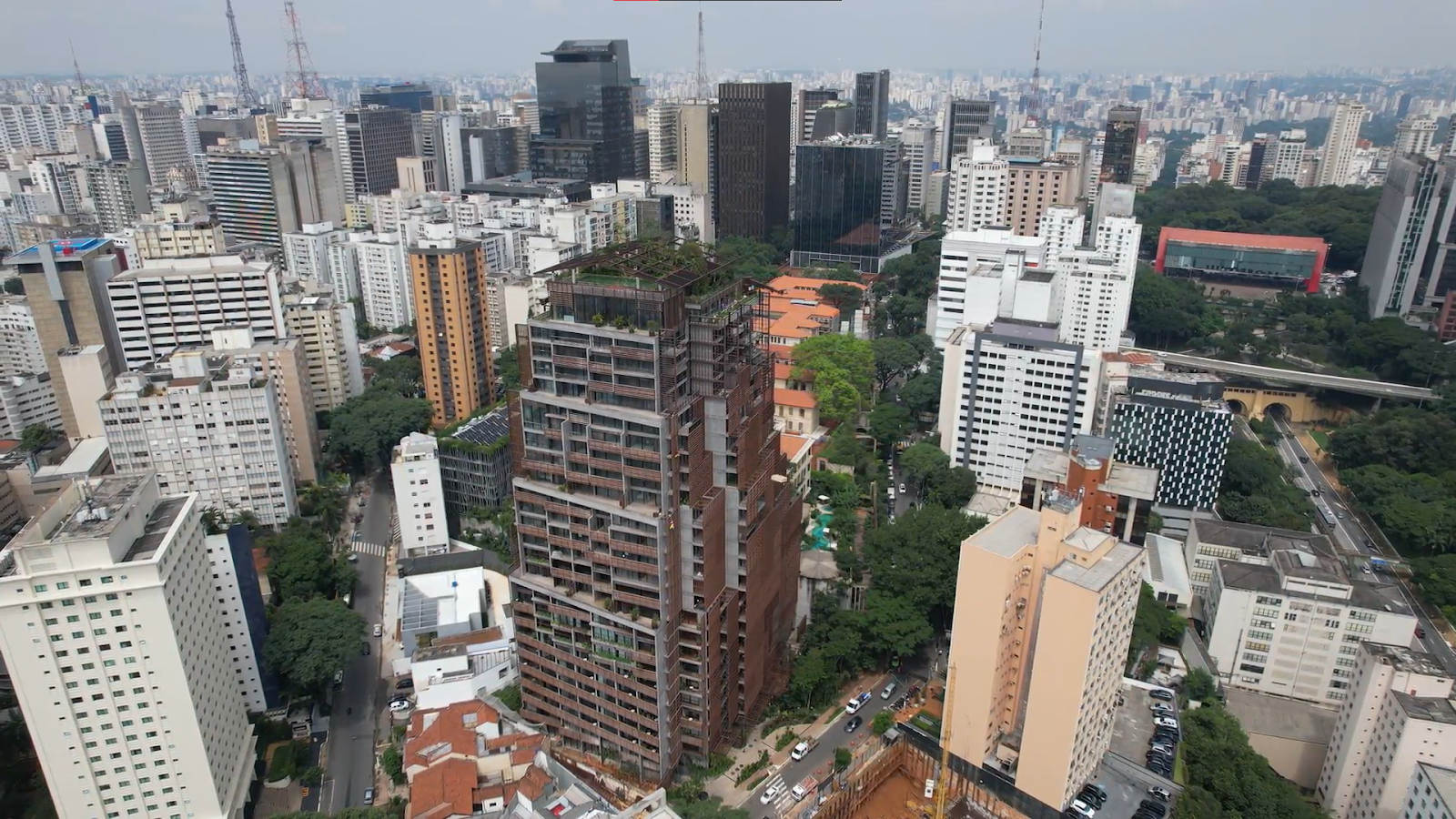 Foto de perspectiva superior da cidade de São Paulo com prédio marrom à frente, outros prédios ao fundo e o Masp no canto superior direito.