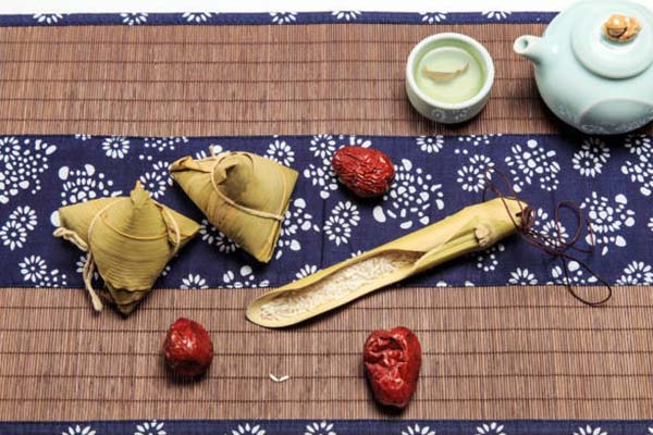 A traditional table setting with zongzi (rice dumplings), a teapot, cup, dried jujubes, and rice grains on a bamboo mat with floral patterns.