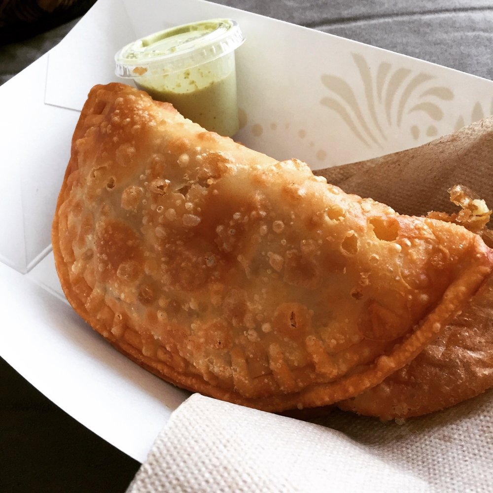 Photo from Caracas Empanadas' Yelp page. A close-up of an perfectly fried, crispy empanada in a disposable food tray. A closed container of a bright green sauce is behind.