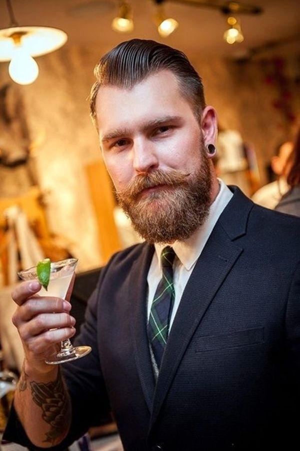 Picture of a man holding a glass and rocking a dapper outfit with his handlebar mustache