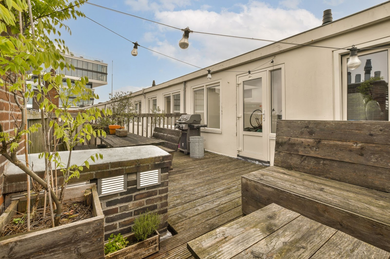 A long wooden deck attached to a white home with wooden tables and chairs.