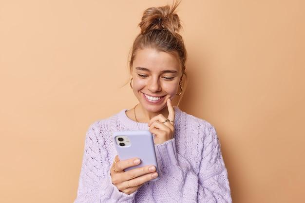 Free photo pleased young woman smiles happily keeps index finger on cheek concentrated into smartphone device reads pleasant message wears casual jumper isolated over beige background. cellular technology