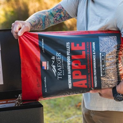 A man with a tattooed arm is pouring Traeger wood pellets into a wood pellet grill. The pellets are apple-flavoured.