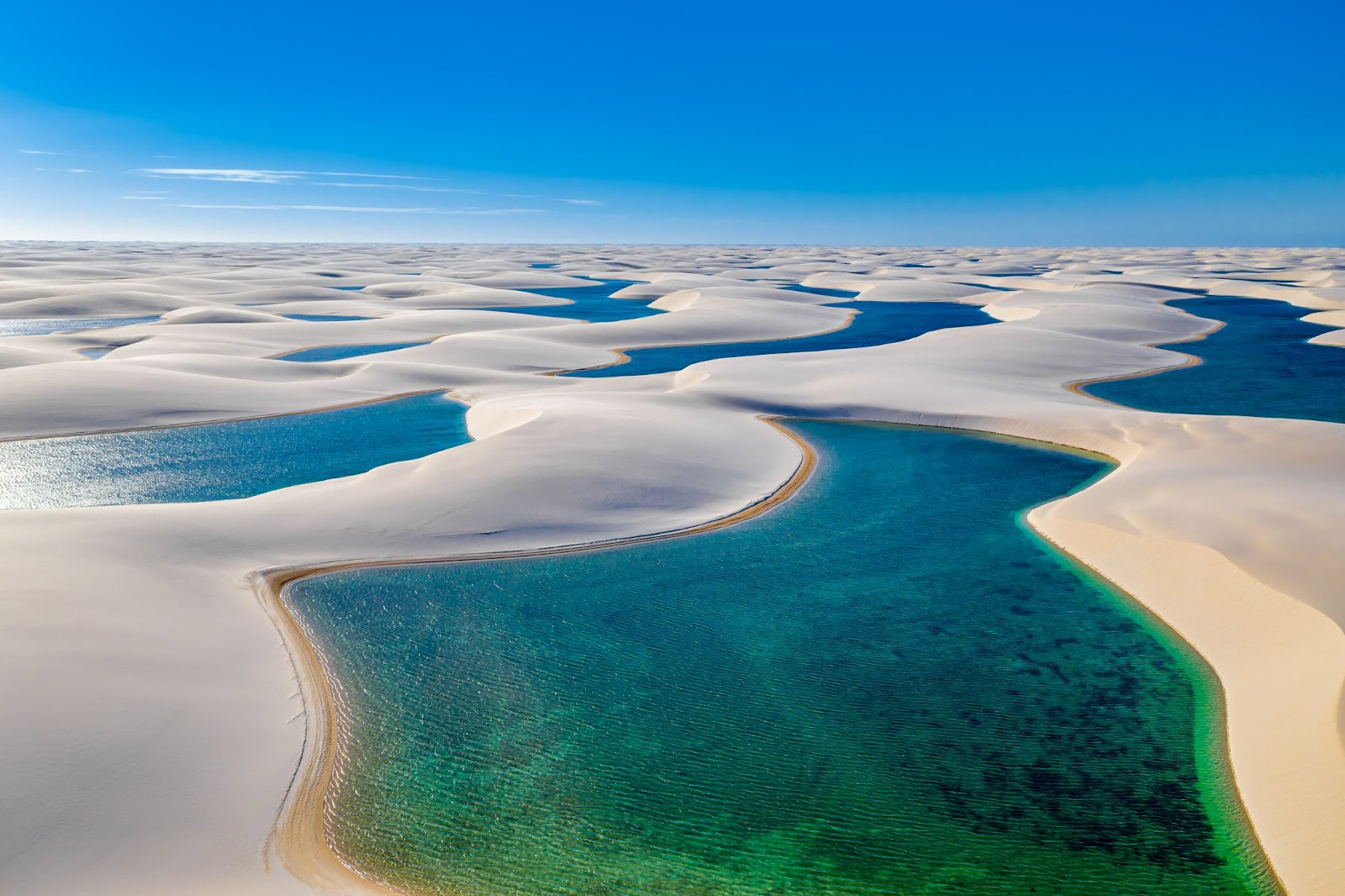 Vista panorâmica dos Lençóis Maranhenses. Grandes dunas de areia branca aparecem intercaladas com lagoas de cor azul intensa. O céu está azul e sem nuvens.