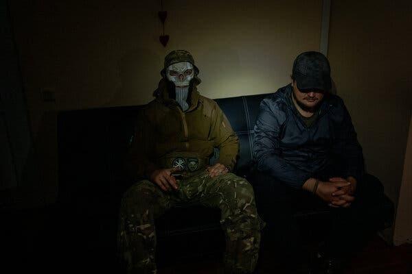 Two men sit side by side in a dimly lighted room, both concealing their faces, one behind a mask and the other under the brim of his cap.