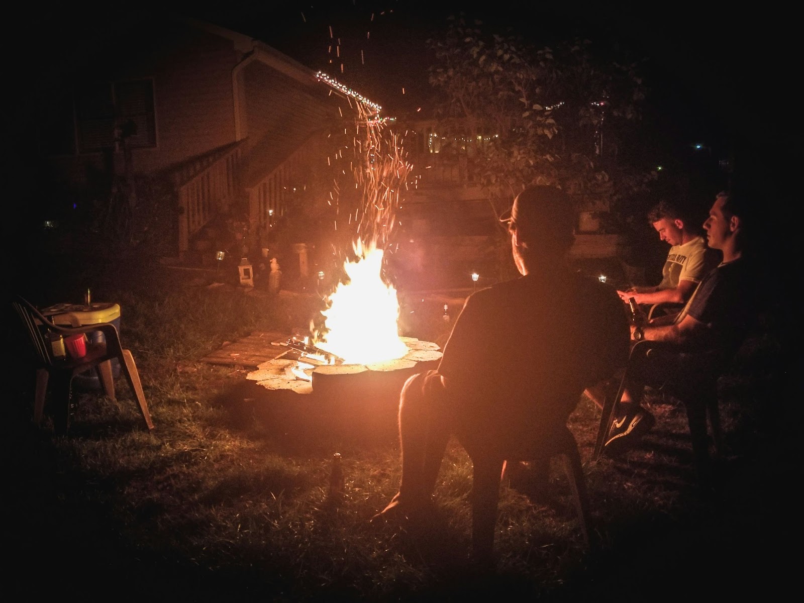 three young men at a cabin sitting having drinks around a wildfire with crackling embers rising into the sky, highlighting potential causes of forest fires in BC