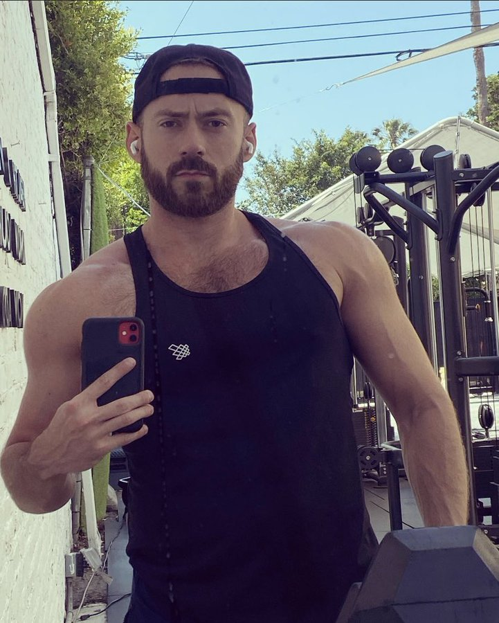 Scott Lazarus taking an iphone mirror selfie in an outdoor gym wearing a black tank top and smirking