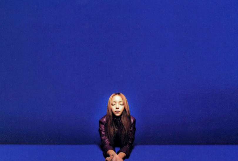 A shot of Namie Amuro from the Genius 2000 album shoot. Namie is wearing a maroon leather jacket, leaning on a blue surface, with a blue backdrop behind her. The same setup for the Genius 2000 album cover, minus the butterflies. (Image credit: Namie Amuro Toi et Moi V4 @ https://www.amuro.fr/)