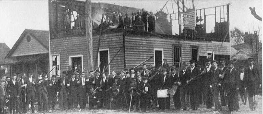 A large group of white men in suits and hats stand in front of a destroyed two-story building.  The roof and second floor are gone and smoke is still coming from the beams.  Many of the men are holding guns.  At least one child is seen at the front of the picture. 