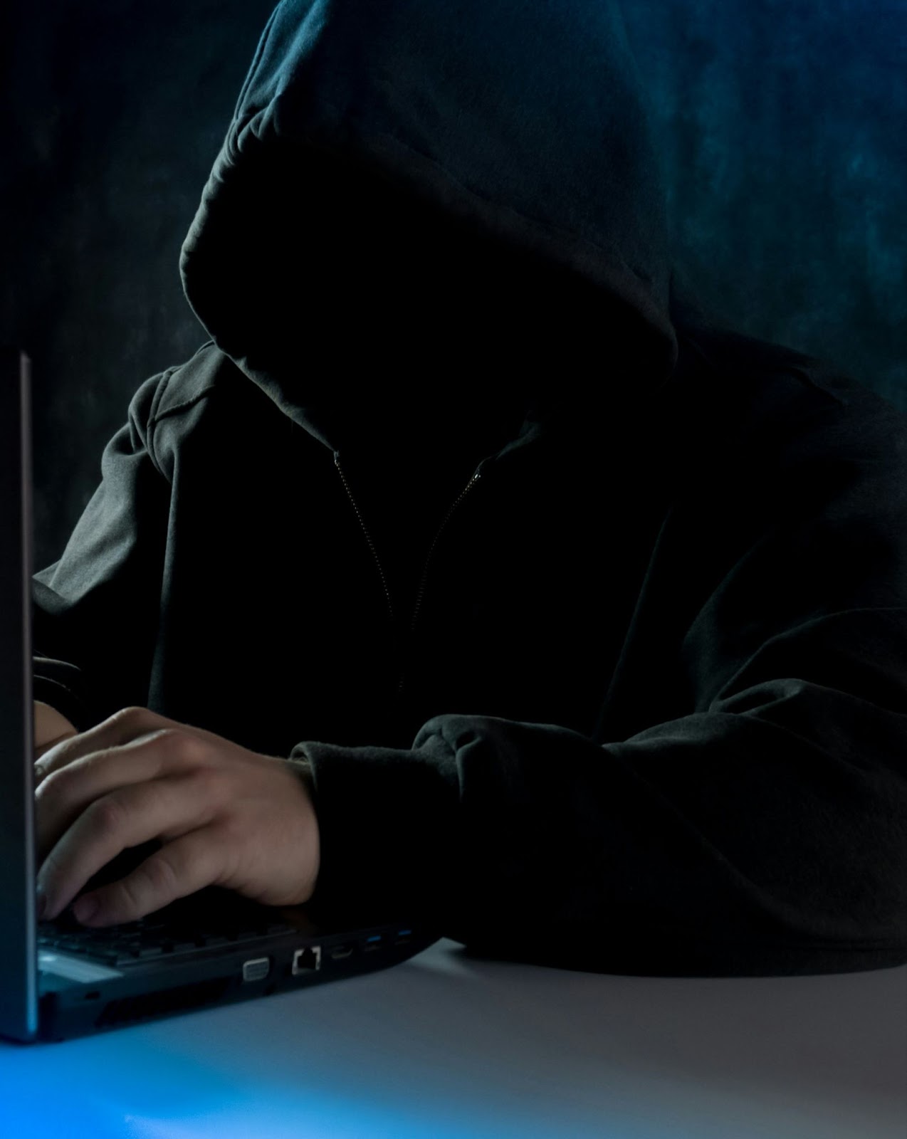 A shadowy figure wearing a black hoodie is typing on a laptop.