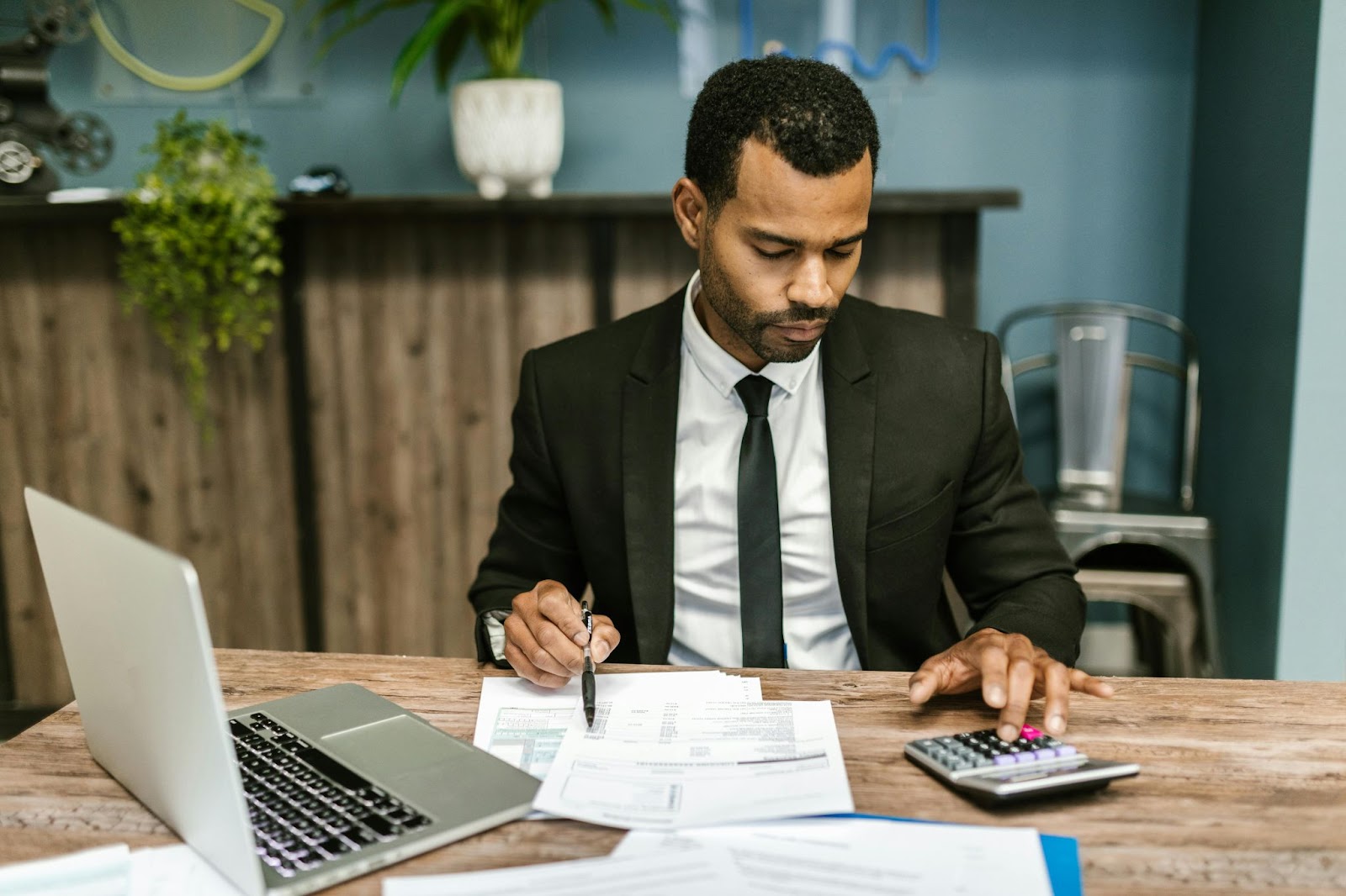 A man in a suit works at a desk with financial documents, a calculator, and a laptop.