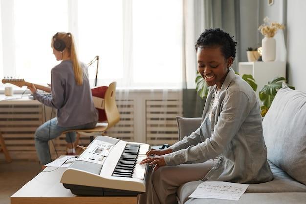 Side view portrait of smiling black woman playing synthesizer at home and composing music in studio