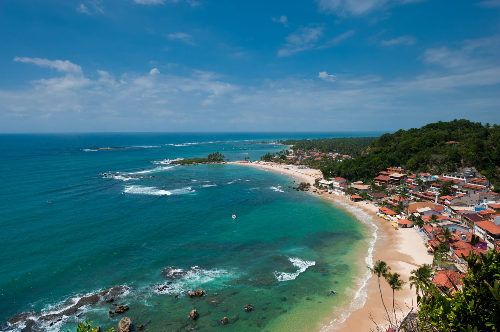 Vista aérea da Primeira, Segunda e Terceira Praia do Morro de São Paulo. Mar azul cristalino ao lado esquerdo da imagem, em frente à longa faixa de areia que leva até as casa e estabelecimentos locais.