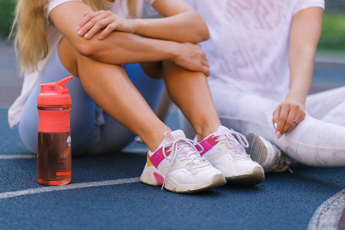 Free Crop anonymous female friends in activewear sitting on sports ground after training near bottle with water in daytime Stock Photo