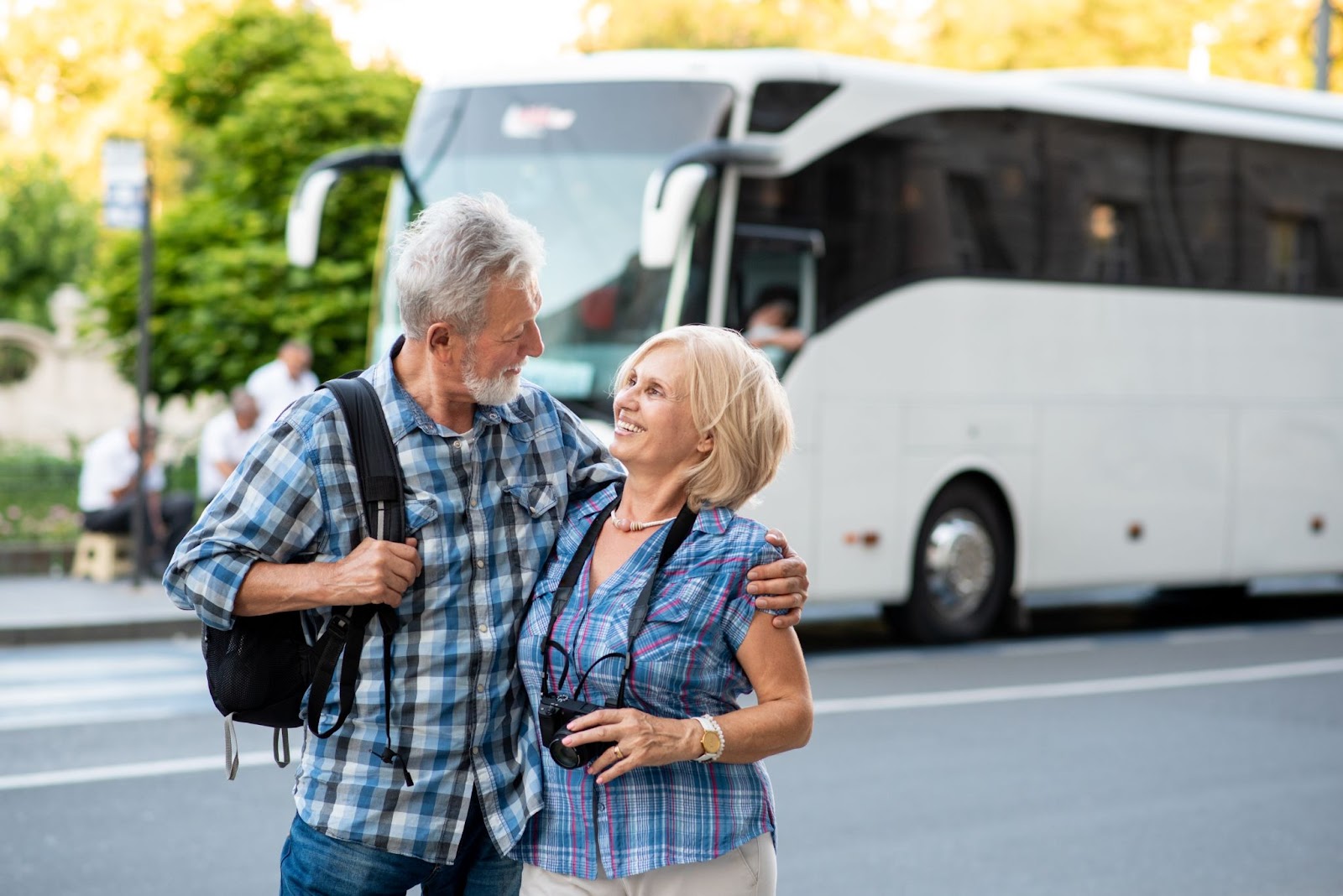 Homem e mulher que parecem ter entre 60 e 70 anos. Eles carregam mochilas e câmeras fotográficas e sorriem um para o outro, abraçados. Ao fundo, aparece um ônibus de viagem branco.