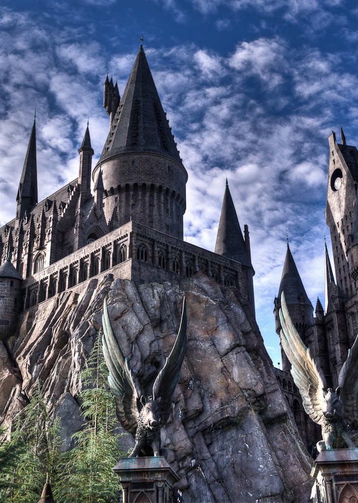 A stone castle with turrents and walls sitting on top of a cliff. In the foreground there are stone two winged-boars on pillars. This is the Hogwarts Castle in Harry Potter World at Universal Studios in Florida. 