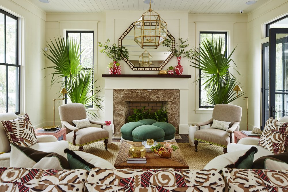 Eclectic living room vignette with in a windowed room with fireplace. The room has a patterned sofa, octagonal mirror above the mantle, tropical potted plants flanking the fireplace, emerald ottoman, and red decorative vases.