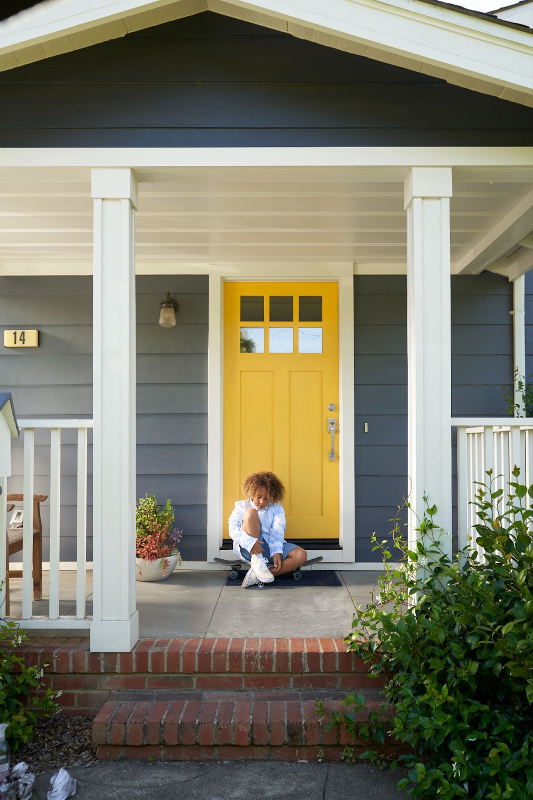 The front of a grey house with a vibrant yellow front door and a kid sitting on the porch.