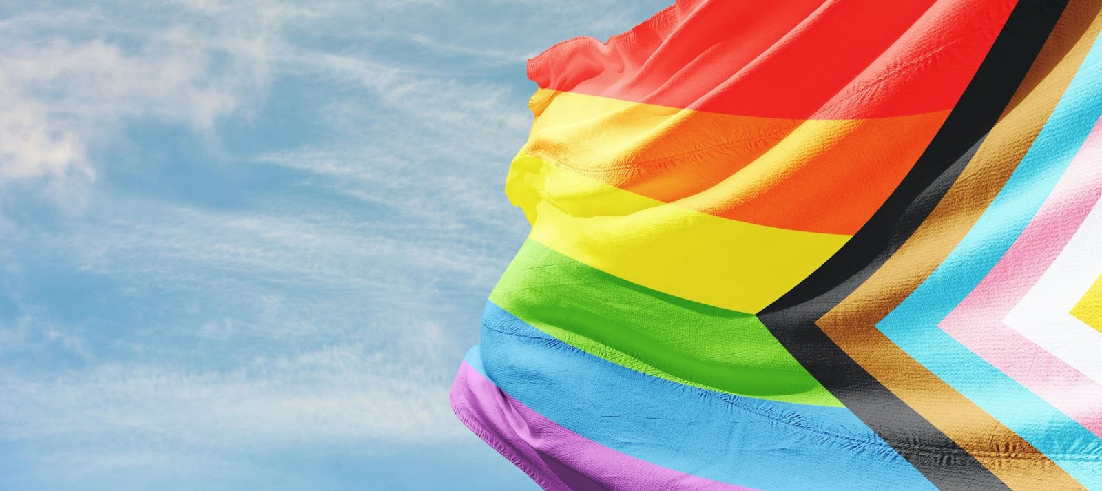 Bandeira representativa do movimento LGBTQIAPN+, com as cores do arco-íris e mais, balançando ao vento e revelando um céu azul claro com nuvens brancas ao fundo.