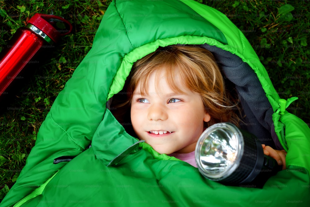 Kleines Vorschulmädchen im Schlafsack beim Camping. Outdoor-Aktivitäten mit Kindern im Sommer. Spaß- und Abenteuercamp, Urlaub mit Familie und Freunden oder Wochenendausflug. Porträt eines Kindes mit Taschenlampe