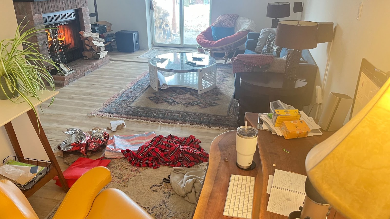 A lived-in room with a variety of items scattered around, suggesting everyday use or recent activity. On the left, there's a fireplace with a visible flame behind a mesh screen, flanked by firewood and a plant on a wooden shelf. The floor is covered with a huge area rug over which a smaller rug is laid. In the center, a round glass coffee table is surrounded by a cozy orange papasan, a teal sofa with throw pillows, and a side table with lamps. Strewn across the room: a red and black plaid blanket, a grey hoodie, foil wrappers, papers, and a couple of boxes with bright yellow packaging. On the right, there's a desk with a keyboard, a notepad with doodles, and a white tumbler. The room has a cozy ambiance, accentuated by the natural light coming through the glass patio door on the left. The scene captures a casual, personal space with signs of daily life and work.