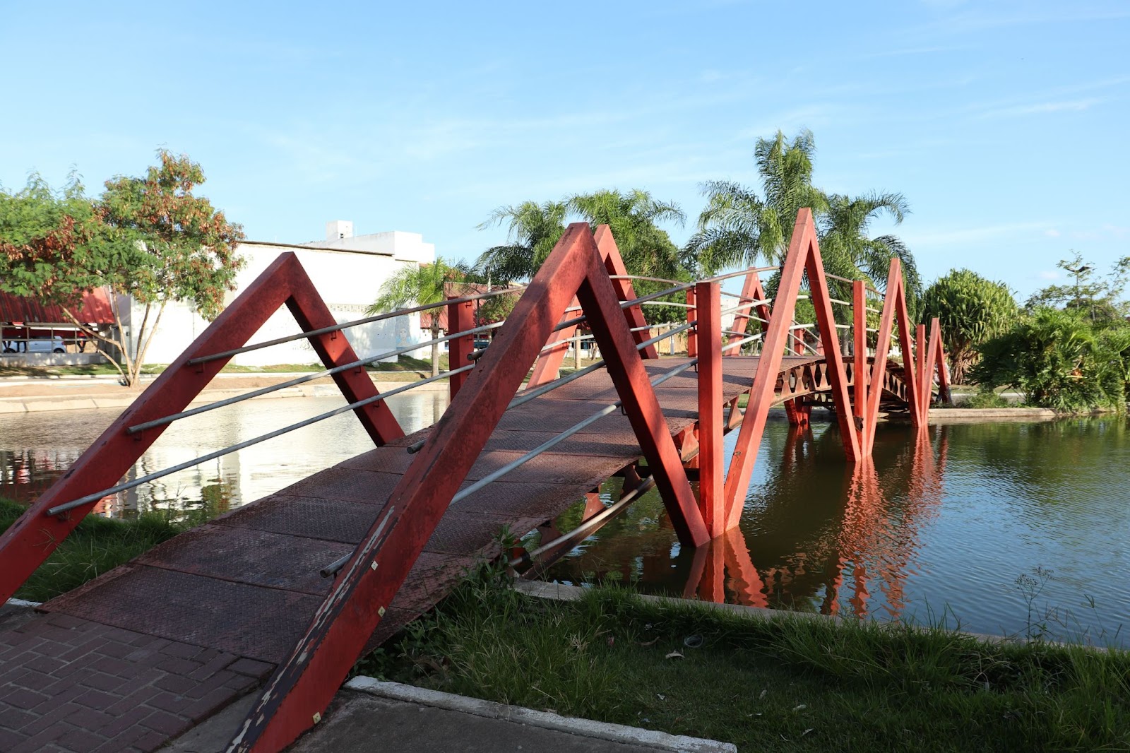 Ponte vermelha sobre o lago do Parque Ceci Cunha. Água verde, com árvores do outro lado da ponte