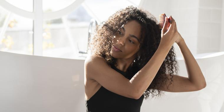 Imagem mostra uma mulher de cabelos cacheados massageando os cabelos, dentro de um quarto com fundo claro.