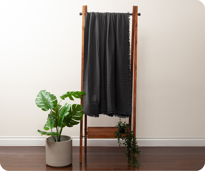 Our charcoal grey Muslin Gauze Blanket in Obsidian shown hanging on a wooden rack in a room.