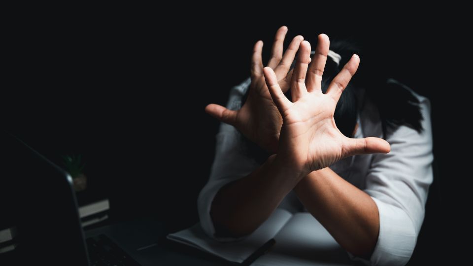 A person in a white shirt extending their hand forward in a defensive or protective gesture, with a dimly lit workspace in the background