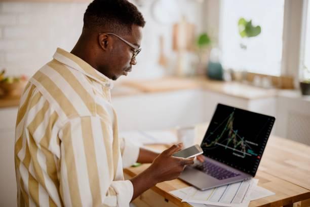 Young man investing or trading in Bitcoin or other cryptocurrencies Young African man working with blockchain technologies. Man analyzing crypto graph on laptop screen. Bitcoin / Cryptocurrency concept. forex trading stock pictures, royalty-free photos & images