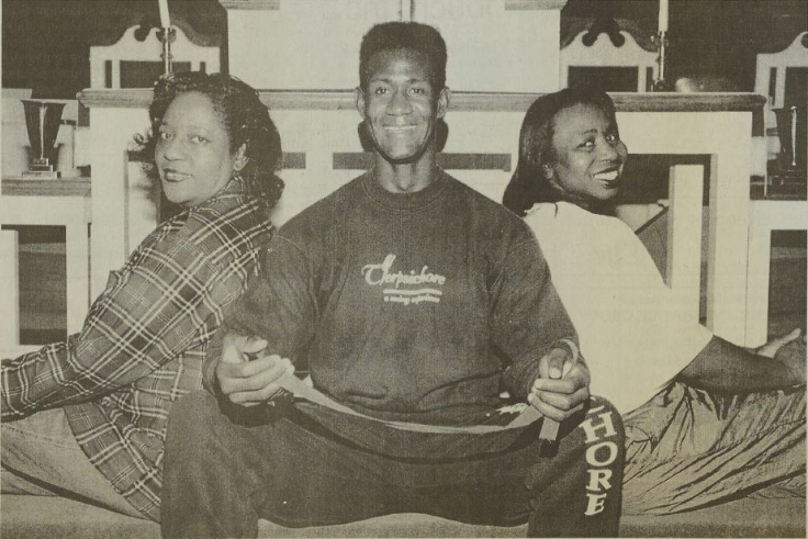 Tomlinson seated between two compatriot dancers. He is wearing a sweatsuit and holding a ribbon in his hand.