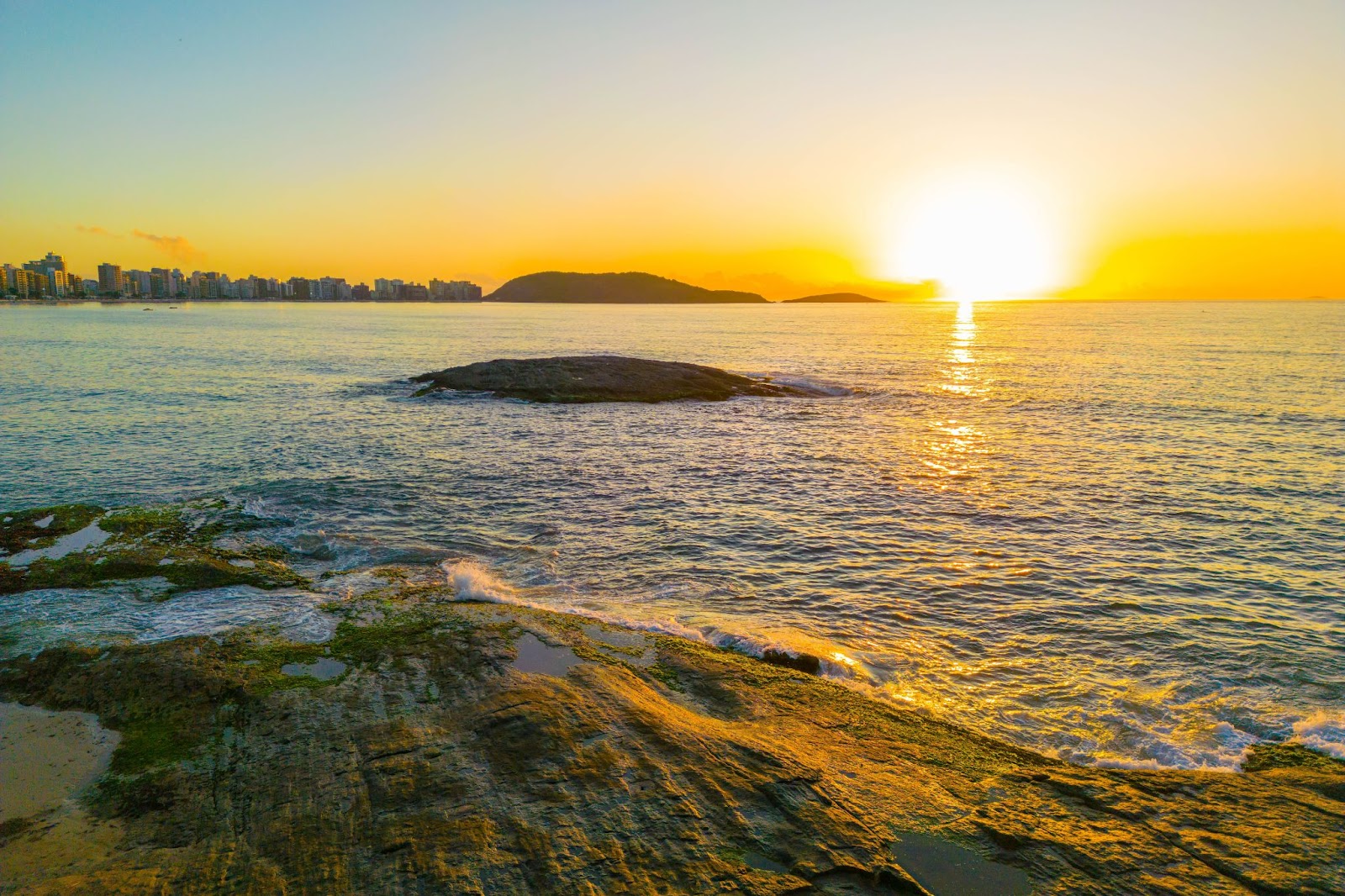 Nascer do sol na Praia do Morro. Mar sem ondas com pequeno morro rochoso, refletindo o amarelo alaranjado do sol.