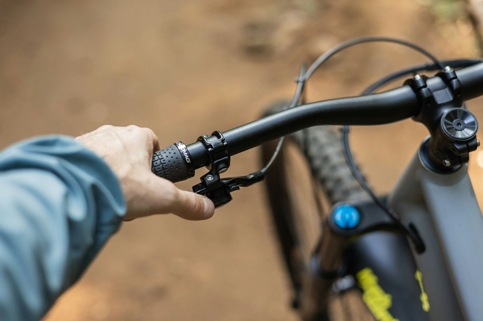 A hand holding the bars of a mountain bike against a dirt ground background. Their thumb is on the gear shift. 