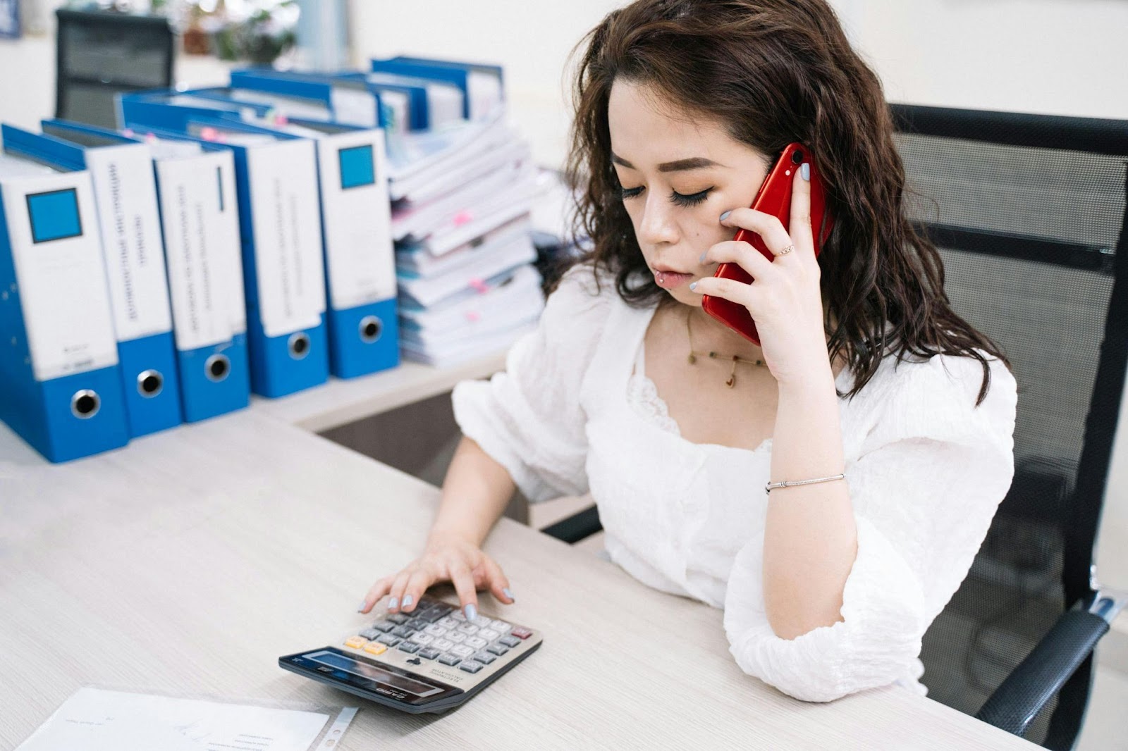 A focused woman talking on a red phone while calculating something with a calculator at a desk with binders in the background