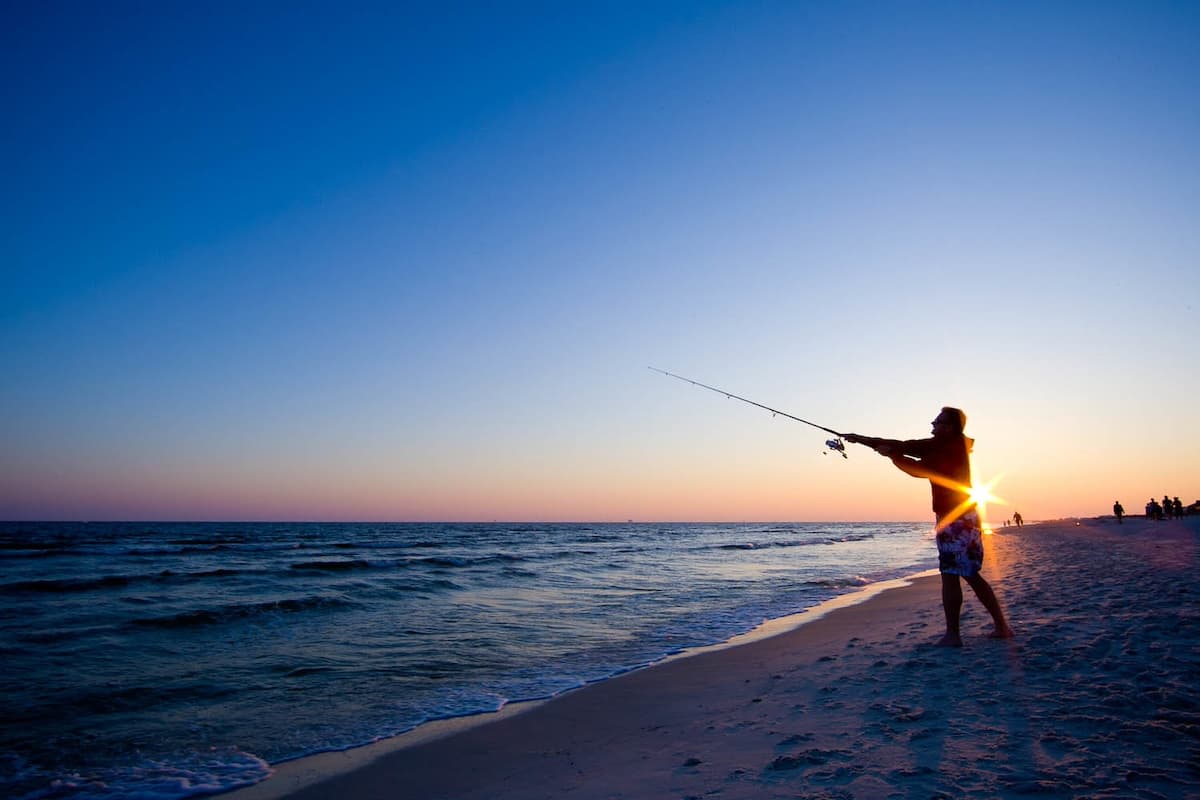 Man surf fishing on the waters edge at Fort Morgan Beach with sun setting in background