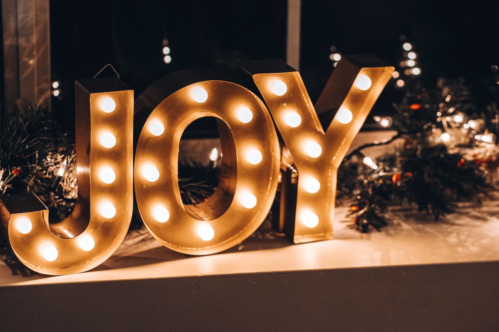 A metal-framed light display spelling JOY is sitting on a counter with various Christmas-themed greenery.