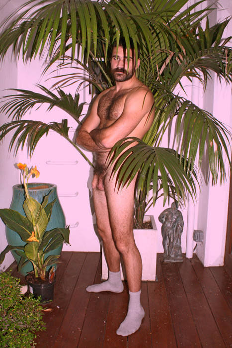 gay male poses behind houseplant palm in nothing but his gym socks while looking to the camera with his arms crossed and showing off his flaccid cock